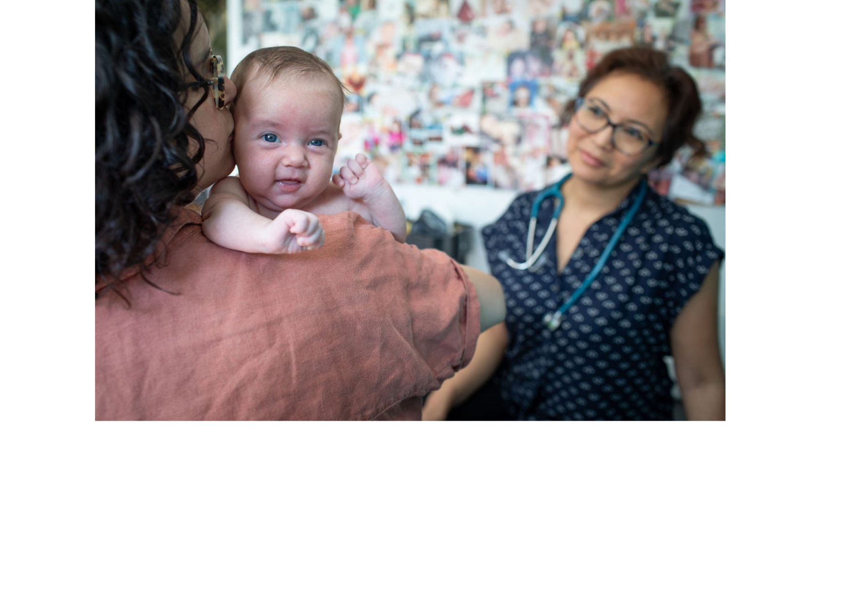 A mother kisses her infant with midwife seated in background in front of a large photo wall of past clients' images.