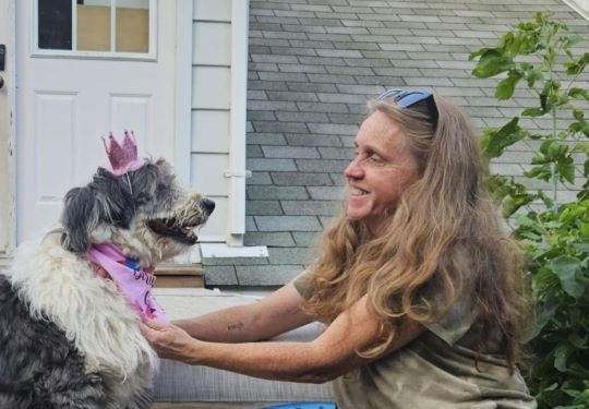 Celeste sits on a parch smiling warmly at a dog wearing a pink tiara.